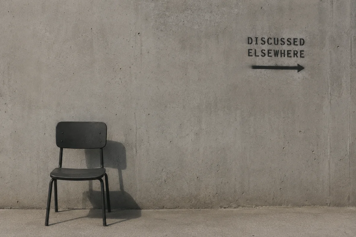 A lone chair sits against a bare concrete wall bearing the stencilled words DISCUSSED ELSEWHERE with an arrow pointing right