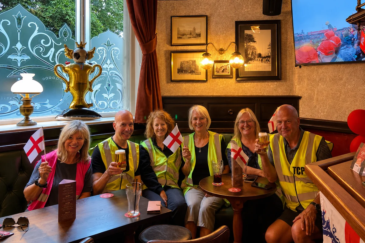 Tottington Civic Pride volunteers in high-visibility vests raising a glass in a village pub, holding St George&rsquo;s Cross flags, with a golden World Cup trophy balloon in the window behind them.